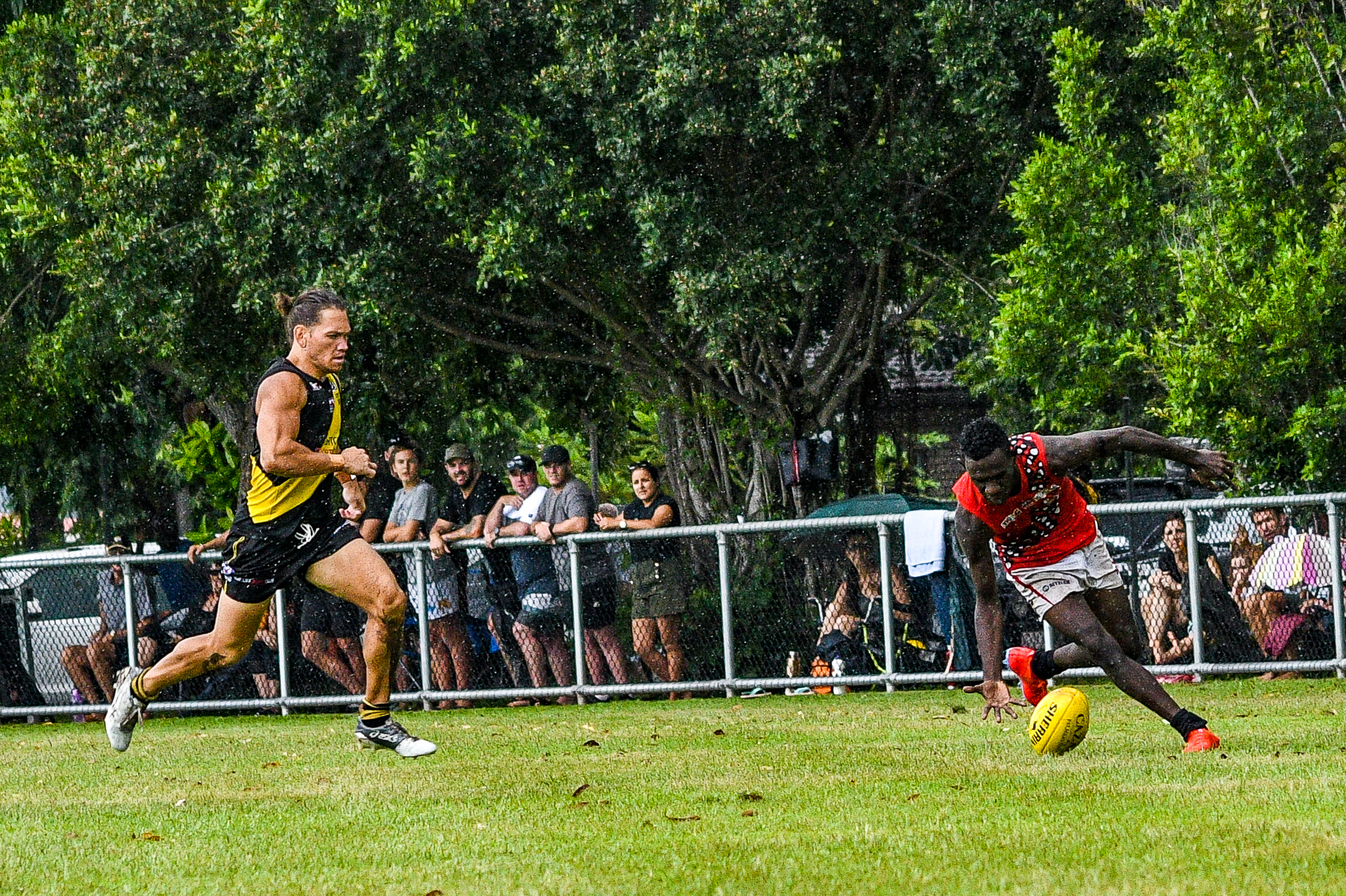 Dion Munkara stoops to pick up a wet ball… Tiwi Bombers v Nightcliff Tigers, NTFL round 15, January 27, 2019.