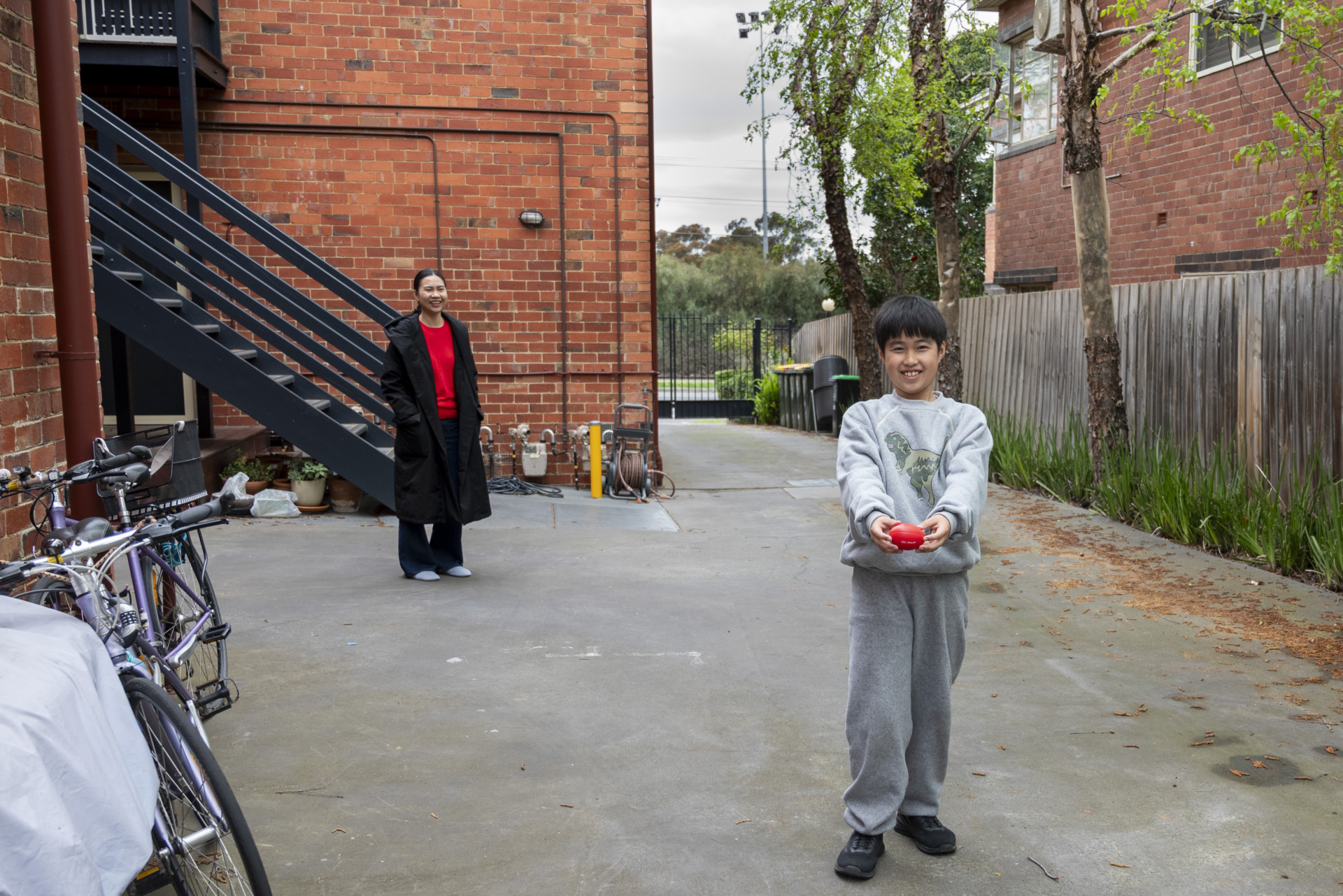 Ling and her son Edward spend time in the backyard of their rented apartment in Elwood. Edward holds a red ball with a cheerful smile while Ling stands in the background, watching him with a warm expression.