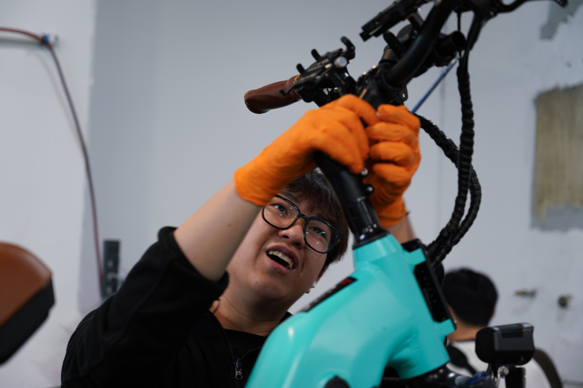 Yang Xihan shows off his e-bike repair skills in the West Melbourne workshop on October 2, 2024. His orange work gloves and cheerful expression reflect his enthusiasm for the job. &ldquo;It was fate that brought me to this e-bike shop,&rdquo; Yang says, explaining how he found the workshop through social media while searching for a rental bike.
