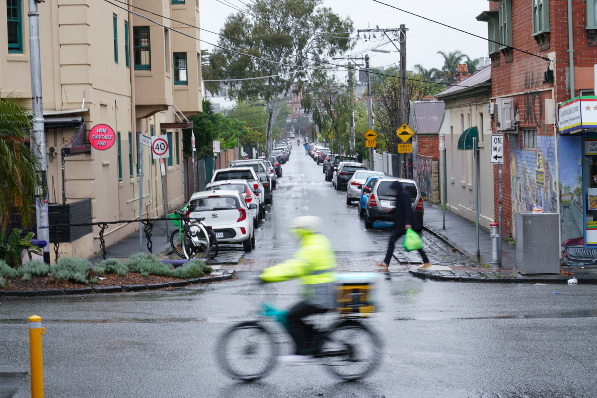 Yang rides his delivery bike down a wet street in South Yarra on September 29, 2024. His bright yellow jacket catches the eye as he moves past old brick houses and parked cars. Someone with groceries crosses the street as Yang heads to his next delivery.